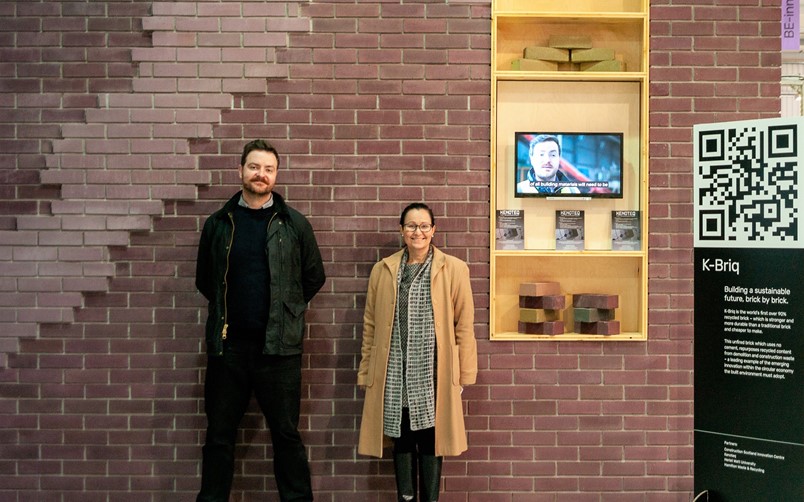 Two creators of K-briq at COP26 in front of wall made from invention